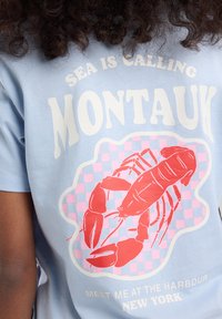 T-shirt in cotone azzurro chiaro con una grafica di un'aragosta su uno sfondo a quadretti rossi e rosa. Presenta il testo "Il Mare Chiama Montauk" sopra e "Incontrami al Porto di New York" sotto.