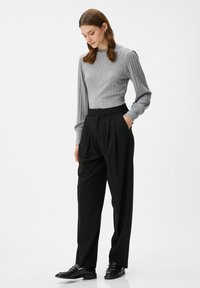 Vrouw in een grijze geribde trui, zwarte high-waisted plooibroek en zwarte instappers, staand met één hand in haar zak, terwijl ze naar beneden kijkt.