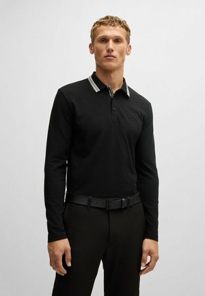 Camisa polo de manga larga negra con un cuello a rayas, que cuenta con una tapeta de tres botones y un logotipo bordado en el pecho. Metida dentro de unos pantalones negros.
