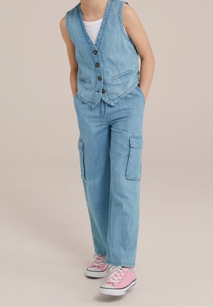 Kind draagt lichtblauw denimvest en cargobroek met roze sneakers, staat met handen in zakken tegen een effen achtergrond.