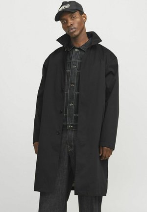 Manteau trench noir oversize avec un col haut et une fermeture à boutons, fabriqué à partir d'un tissu lisse. Porté par-dessus une tenue en denim.