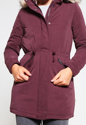 Parka bordeaux avec capuche en fausse fourrure, fermeture éclair frontale et taille avec cordon de serrage. Comprend deux poches latérales zippées et manches longues.