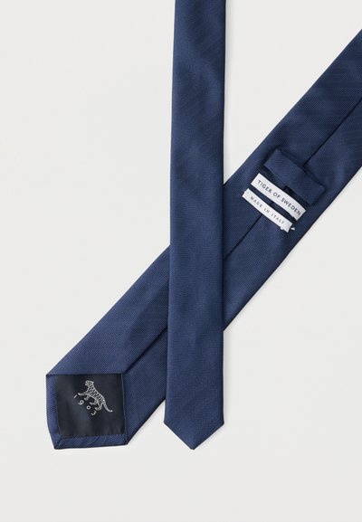 Στενή γραβάτα σε σκούρο μπλε χρώμα με σχέδιο ψαροκόκκαλου, με την ετικέτα «Tiger of Sweden» και «Made in Italy», απλωμένη επίπεδη σε λευκή επιφάνεια.