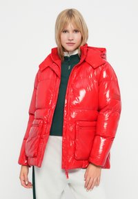 Veste rembourrée rouge et brillante avec capuche amovible, poches avant et fermeture éclair. Portée sur un haut vert et un pantalon gris clair.