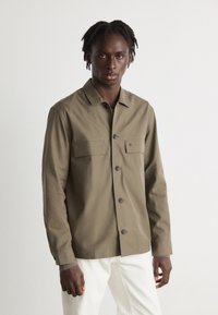 Olive green button-up shirt with a collar, chest pockets, and black buttons. Long sleeves feature buttoned cuffs. Worn over white pants.