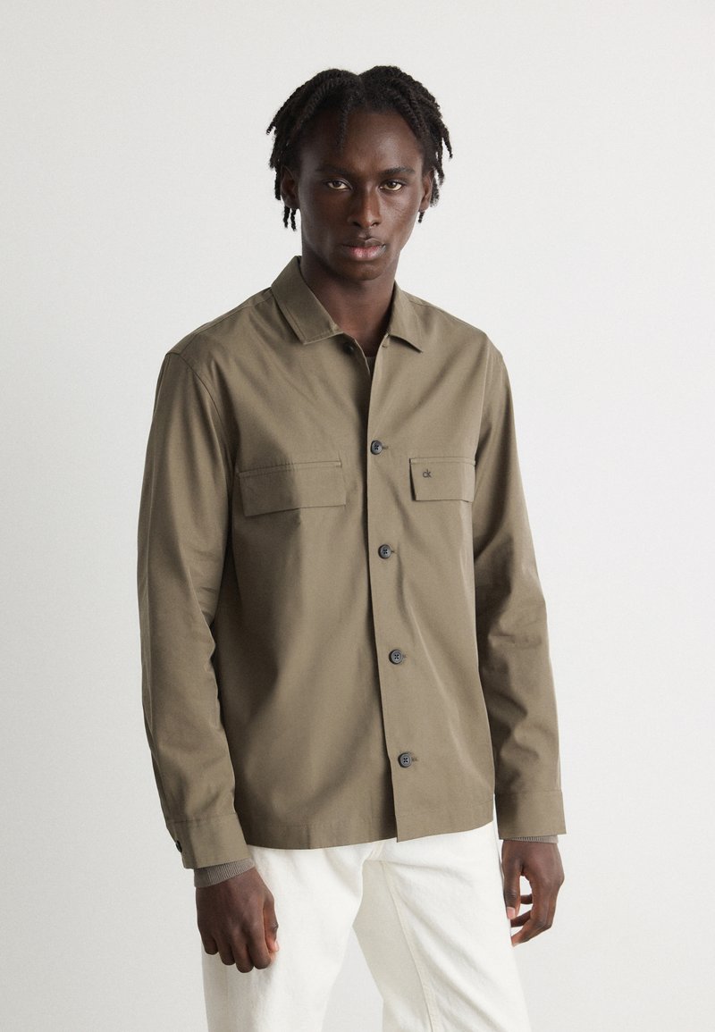 Olive green button-up shirt with a collar, chest pockets, and black buttons. Long sleeves feature buttoned cuffs. Worn over white pants.