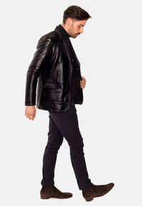 Chaqueta de cuero negra con un corte a medida, que presenta dos botones y bolsillos laterales, llevada sobre un suéter de cuello alto oscuro y combinada con pantalones oscuros y botas.