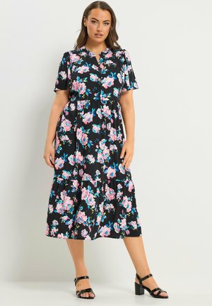 Kvinde iført en sort midi kjole med pink og blå blomsterprint og korte ærmer, parret med sorte sandaletter med hæl, står op ad en hvid væg.