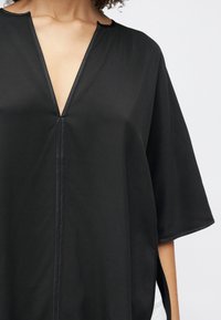 Sort oversized bluse med V-udskæring, korte ærmer og sidenedfældninger. Fremstillet af glat stof med synlige sømme langs halsudskæringen.