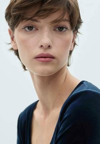 Portret van een persoon met kort, bruin haar, blauwe ogen en natuurlijke make-up, gekleed in een diepblauwe fluwelen top met een brede halslijn.