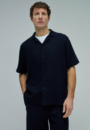 Joven hombre de pie, con una camisa oscura de manga corta abotonada sobre una camiseta blanca, con las manos parcialmente en los bolsillos del pantalón.