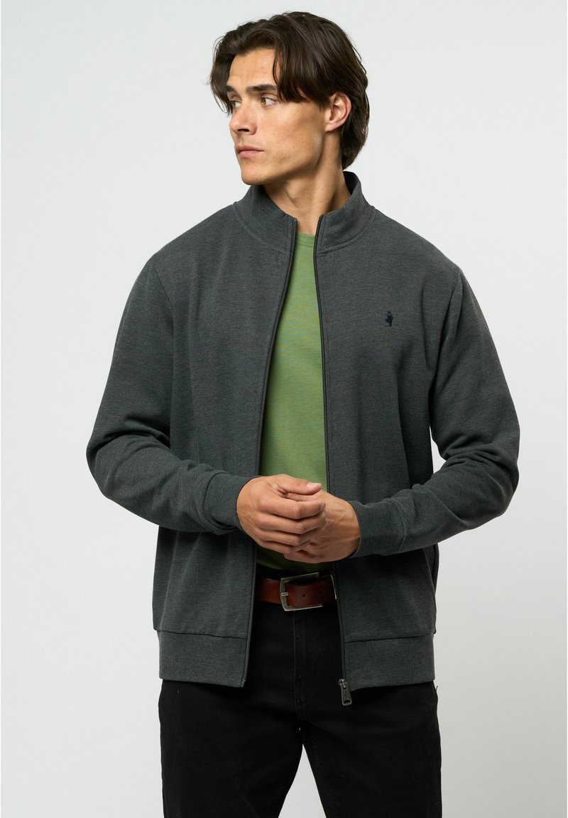 Chaqueta de cremallera gris oscuro en tela suave, con cuello alto, puños acanalados y un sutil acento de logo en el pecho. Llevada sobre una blusa verde.