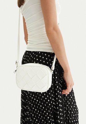 Woman wearing white ruched top and black pleated skirt with white polka dots, carrying a small white quilted crossbody bag.