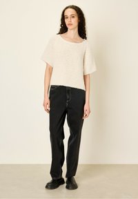Top en tricot crème à manches courtes avec un motif texturé, associé à un jean noir ample et des chaussures noires épaisses. Fond lisse.