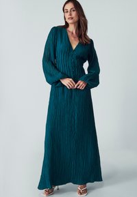 Tyrkysové šaty s dlouhým rukávem a texturovanou tkaninou, zapínání na knoflíky vpředu a splývavý střih. Mají široké rukávy a výstřih do V.