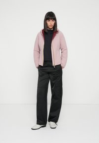 Light pink zip-up jacket with a maroon hood, black high-neck top, black cargo pants, and white ankle boots in a plain background.