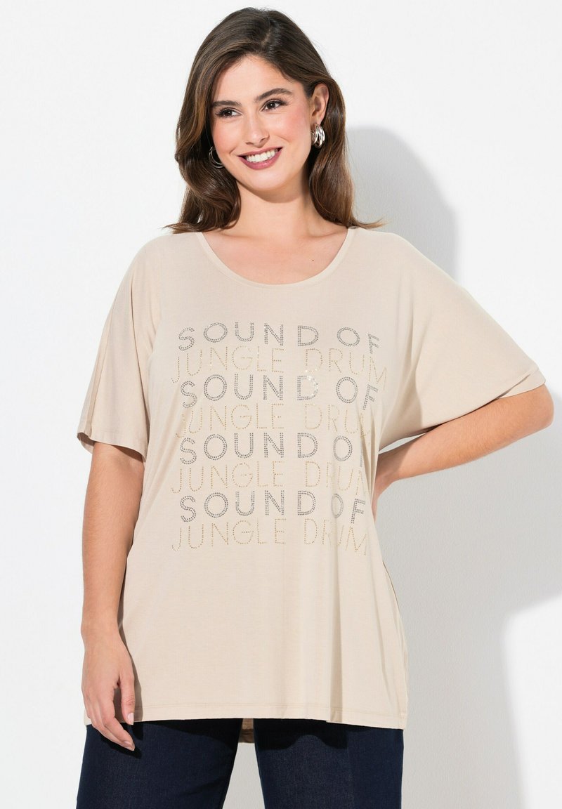 Beige oversized t-shirt med løse ærmer, prydet med trykt tekst "SOUND OF JUNGLE DRUM" i skinnende accenter. Kombineret med mørke jeans.