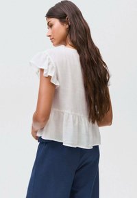 Weißes, leichtes, kurzärmliges Oberteil mit Rüschensäumen und einem Peplum-Saum. Getragen mit marineblauen Hosen, von hinten betrachtet.