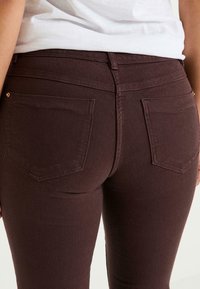 Jean skinny bordeaux avec une texture lisse, comportant deux poches arrière et des passants de ceinture, photographié de la taille vers le bas.