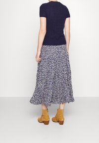 Žena v námořnickém krátkém pleteném topu, modro-bílé květinové midi sukni a béžových semišových kotníkových botách, stojící čelem k nám.