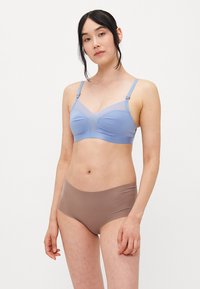 Kvinne som står iført en lyseblå bh med mesh-detaljer og beige sømløst høyhåndet undertøy mot en enkel hvit bakgrunn.