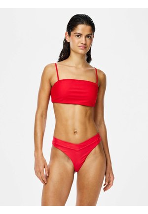 Žena s mokrými vlasmi, ktorá má na sebe červené dvojdielne plavky so rovnakým výstrihom a nohavičkami s vysokým pásom a výstrihom do tvaru V, stojaca na bielom pozadí.