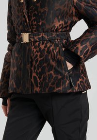 Nahaufnahme einer Person, die eine braun-schwarze Leopardenmusterjacke mit einem Gürtel mit goldener Schnalle und schwarze Hosen trägt, die Hand in der Reißverschlusstasche.