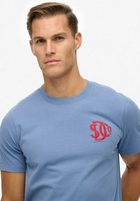 Joven con cabello corto color castaño claro, vistiendo una camiseta de color azul claro con un logo bordado rojo en el pecho, de pie frente a un fondo blanco.