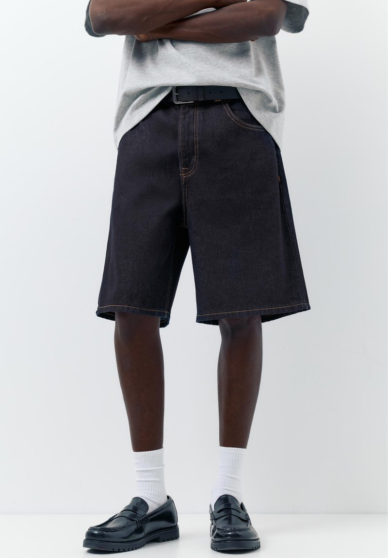 Personne portant un short en jean foncé, un t-shirt gris, des chaussettes blanches montantes et des mocassins en cuir noir, debout devant un fond blanc uni.