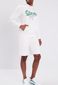 Sudadera blanca con el logo verde "Creeks", capucha con cordón y bolsillo tipo canguro. Combinada con pantalones cortos blancos a juego y zapatillas blancas.