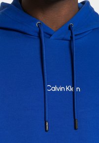Close-up van een blauwe hoodie met trekkoorden en witte "Calvin Klein" tekst op de borst.