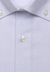 Gros plan sur une chemise bleu clair avec un petit motif en zigzag, présentant un col boutonné et un bouton blanc visible sur la patte de boutonnage.