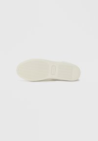 Λευκή σόλα αθλητικού υποδήματος με υφές λαβής και ορθογώνιο λογότυπο ανάγλυφο κοντά στο κέντρο σε λευκό φόντο.