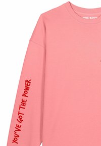 Sweat-shirt rose à manches longues en coton. Il présente le texte "YOU'VE GOT THE POWER" en rouge sur la manche gauche et un petit logo sur la poitrine.