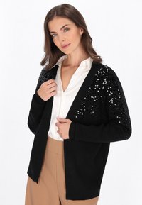 Schwarze Strickjacke mit Pailletten-Schultern, offener Vorderseite, gerippten Manschetten und einer weichen Textur. Tragen über einem weißen Hemd mit Kragen und beigen Hosen.
