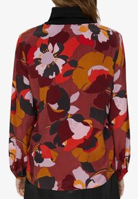 Femme portant un chemisier à manches longues avec un grand imprimé floral abstrait aux tons rouge, orange, noir et rose, vue de dos.