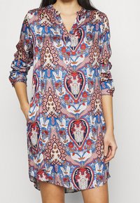 Nainen pukeutuneena värikkääseen pitkähihaiseen tunikamekkoon, jossa on kukkakuvio, sinisiä, punaisia, vaaleanpunaisia ja valkoisia yksityiskohtia sekä napit edessä.