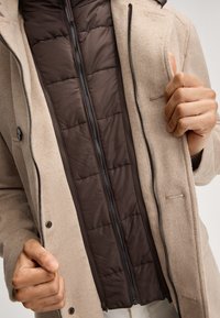 Beige wollen overcoat met knoopsluitingen, voorzien van een bruine gewatteerde voering en een contrasterende rits, die gelaagde texturen benadrukt.