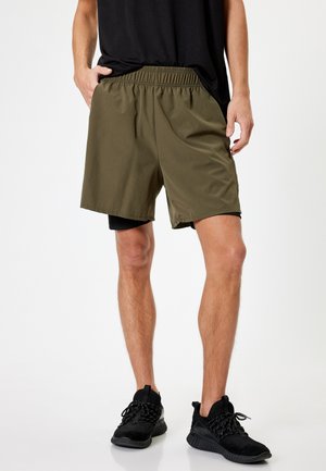 Olivegrønne træningsshorts med elastisk talje, glat tekstur og løs pasform, parret med sorte sneakers.