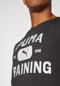 T-shirt en coton noir avec le texte imprimé en blanc "PUMA" et "RAINING" ainsi qu'un logo, design à col rond et usure visible sur l'impression.