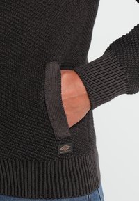 Ciemnoszary sweter wykonany z dzianiny, o teksturze w wzór waflowy, posiada kieszeń boczną oraz ściągacze na mankietach. Metka marki jest widoczna blisko dolnego brzegu.