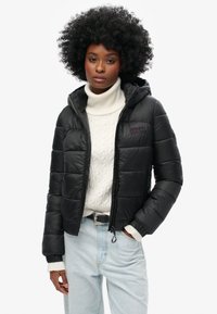 Sort pufferjakke med hætte, der har en lynlåslukning fortil, elastiske manchetter og et subtilt logo på brystet. Bæres over en hvid kabelstrikket sweater.