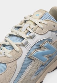Baskets New Balance beige et bleu clair avec panneaux en mesh et en suède, lacets blancs et logo "N" de la marque sur le côté.