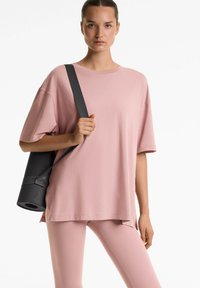 Ylipitkä vaaleanpunainen puuvillapaita lyhyillä hihoilla, yhdistettynä vaaleanpunaisiin leggingseihin. Mallilla on kädessään musta jooga-alusta, joka on kiinnitetty hihnalla.
