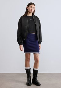 Μαύρο μπουφάν bomber με ριμπ μανσέτες, μαύρο κοντό τοπ, υφασμένη μπλε ναυτικό φούστα με λευκή μπορντούρα και μαύρες μπότες μέχρι το γόνατο με χοντρή σόλα.