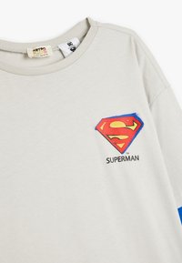 Hellgrau Baumwoll-T-Shirt mit kurzen Ärmeln, das ein rotes und gelbes Superman-Logo auf der Brust und blaue Akzente an den Ärmeln zeigt.