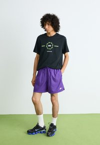 Čierne tričko Nike s zelenou grafickou potlačou, spárované s fialovými šortkami a čiernymi športovými topánkami s modrými akcentmi, na zelenej podlahe.