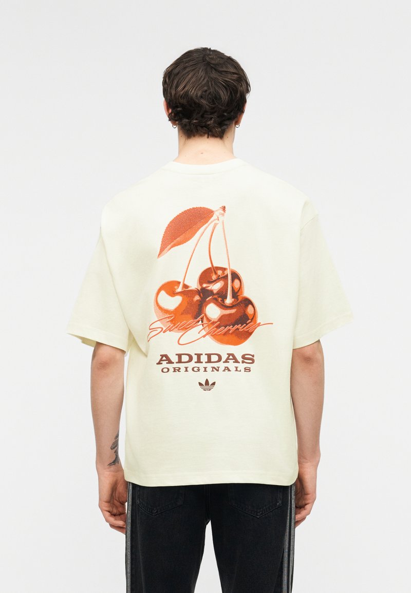Άτομο που φοράει κρεμ T-shirt Adidas Originals με μεγάλο κόκκινο γραφικό κεράσι και κείμενο «Γλυκά Κεράσια» στο πίσω μέρος, στέκεται με την πλάτη γυρισμένη.