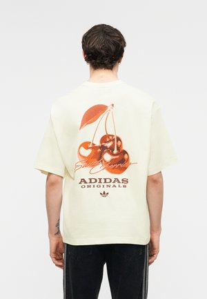 Osoba nosiaca krémové tričko Adidas Originals s veľkou červenou grafikou čerešní a textom „Sweet Cherries“ na chrbte, stojaca otočená chrbtom.