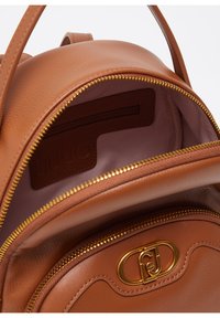 Zaino in pelle marrone con una superficie liscia. Caratterizzato da una cerniera dorata e un'etichetta con logo all'interno. Forma compatta e rotonda con un unico scomparto aperto.